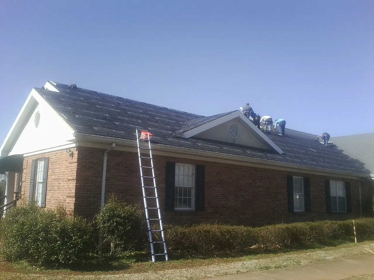 Crew tearing shingles off a brick ranch roof with a ladder set — Metal Roof Repair work by our crew in Chinquapin