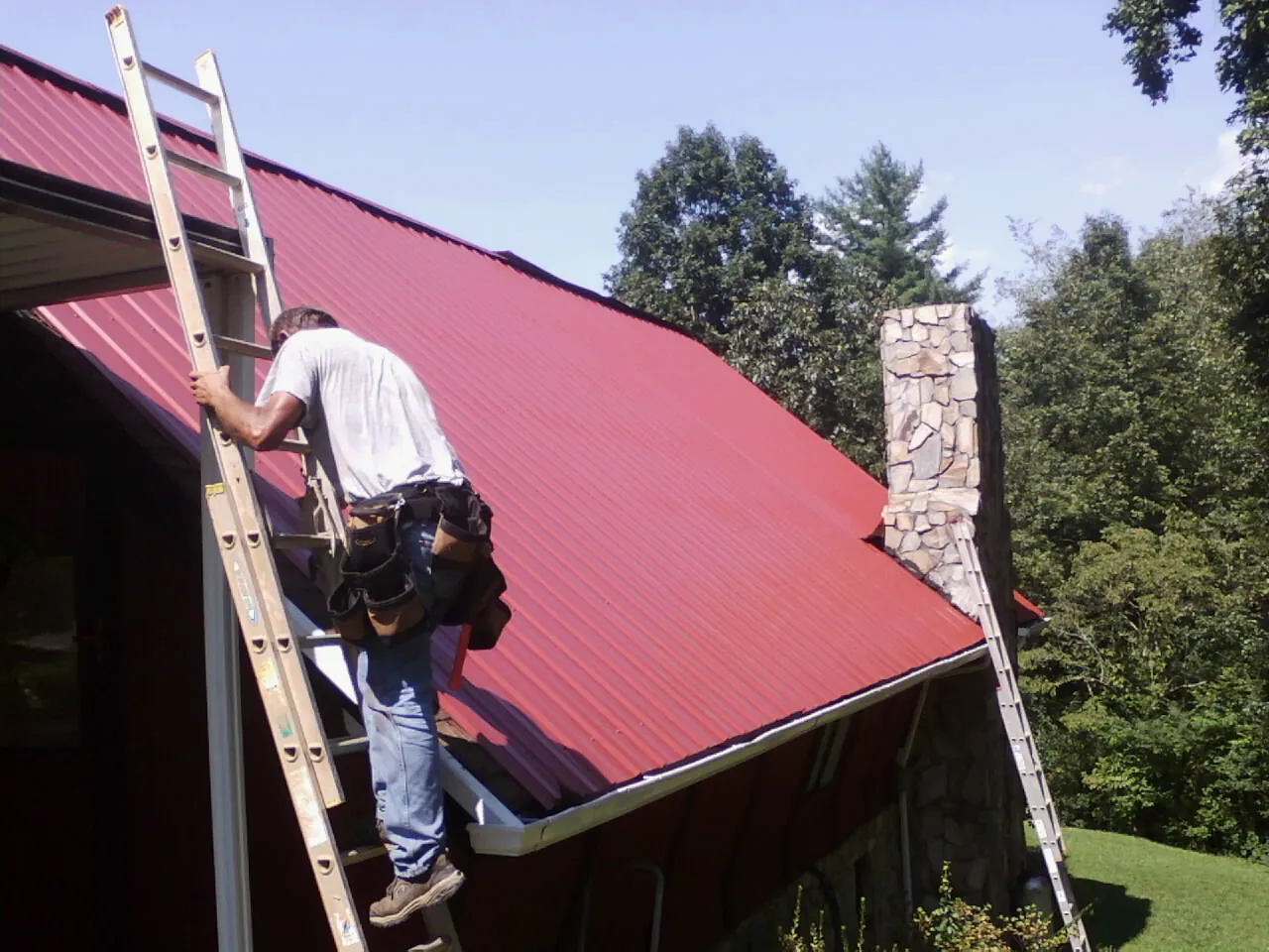 Roofer on a ladder beside a red metal roof and stone chimney — Soffit & Fascia Repair by Roofing Brothers in Chinquapin